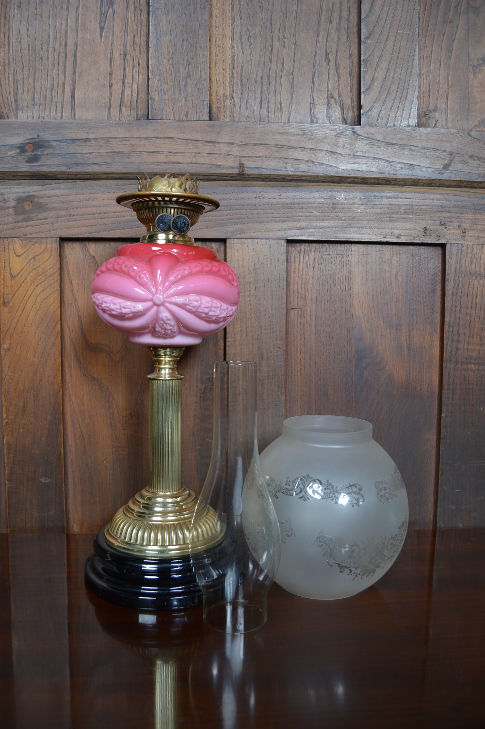A Victorian brass oil lamp with a black base, fluted column, pink patterned glass reservoir, and frosted glass globe shade with etched detail. View IX