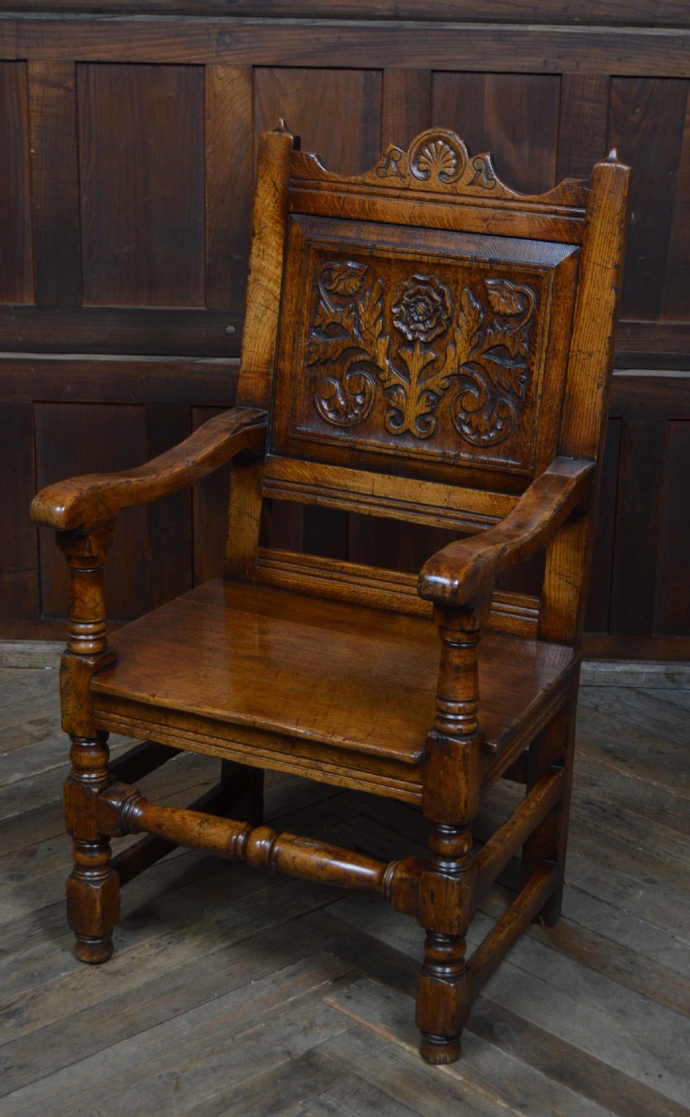 A carved dark oak wainscot style armchair featuring a floral relief panel back, shaped armrests, turned front legs, and wooden stretchers.