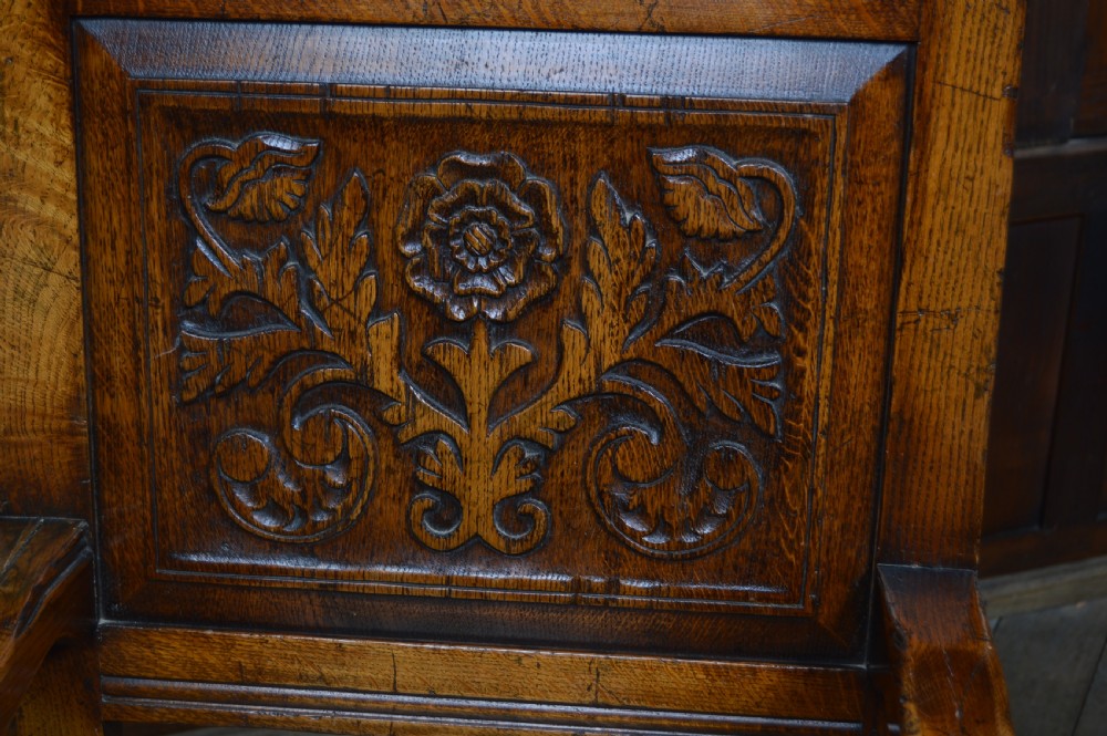 A carved dark oak wainscot style armchair featuring a floral relief panel back, shaped armrests, turned front legs, and wooden stretchers. View IV