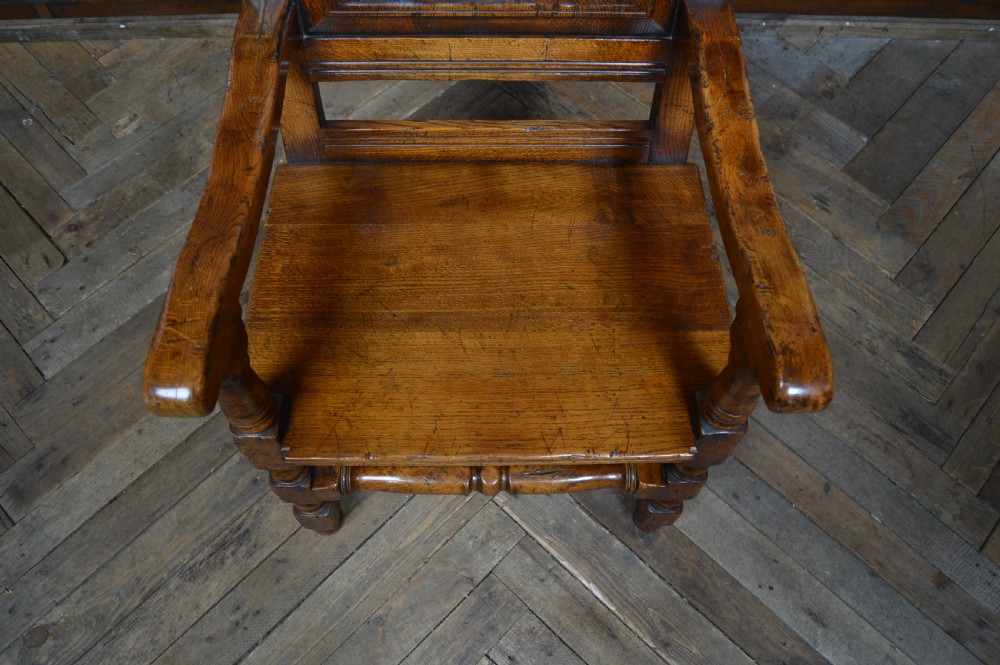 A carved dark oak wainscot style armchair featuring a floral relief panel back, shaped armrests, turned front legs, and wooden stretchers. View V