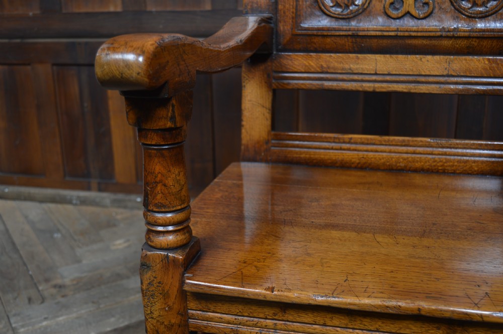 A carved dark oak wainscot style armchair featuring a floral relief panel back, shaped armrests, turned front legs, and wooden stretchers. View VI