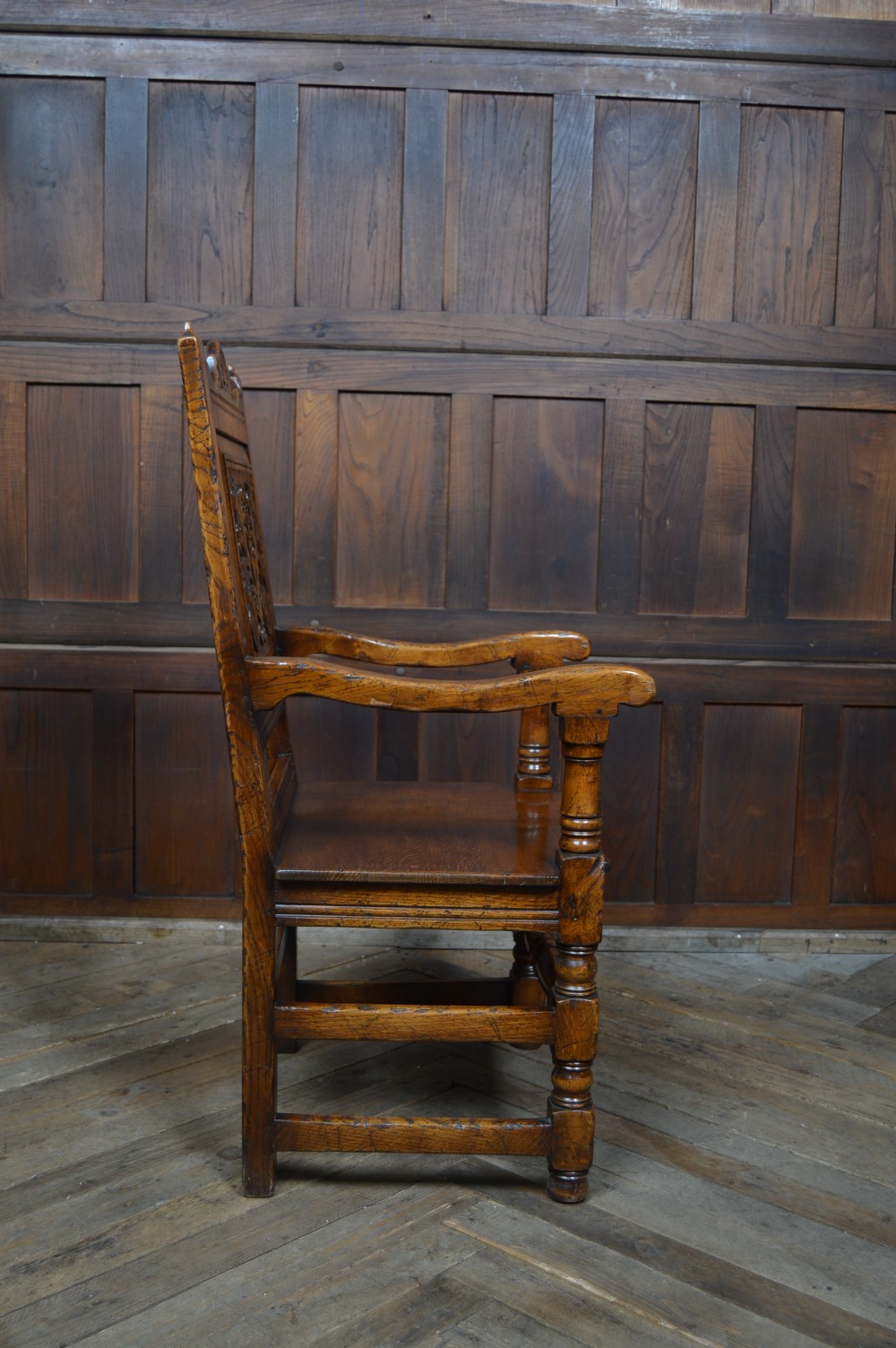 A carved dark oak wainscot style armchair featuring a floral relief panel back, shaped armrests, turned front legs, and wooden stretchers. View IX