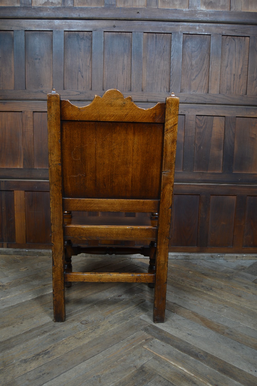 A carved dark oak wainscot style armchair featuring a floral relief panel back, shaped armrests, turned front legs, and wooden stretchers. View X