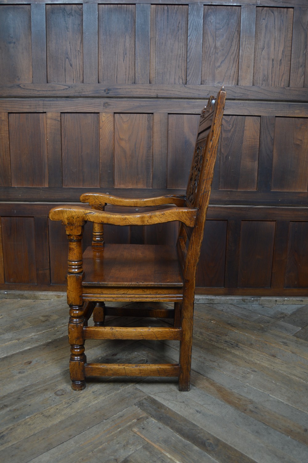 A carved dark oak wainscot style armchair featuring a floral relief panel back, shaped armrests, turned front legs, and wooden stretchers. View XII