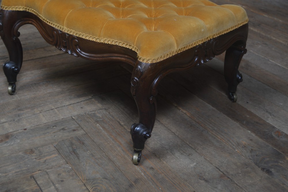 A Victorian low footstool with deep-buttoned mustard yellow velvet upholstery, a carved dark timber frame, and cabriole legs on brass castors. View II