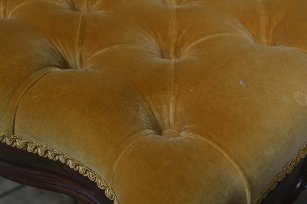 A Victorian low footstool with deep-buttoned mustard yellow velvet upholstery, a carved dark timber frame, and cabriole legs on brass castors. View V