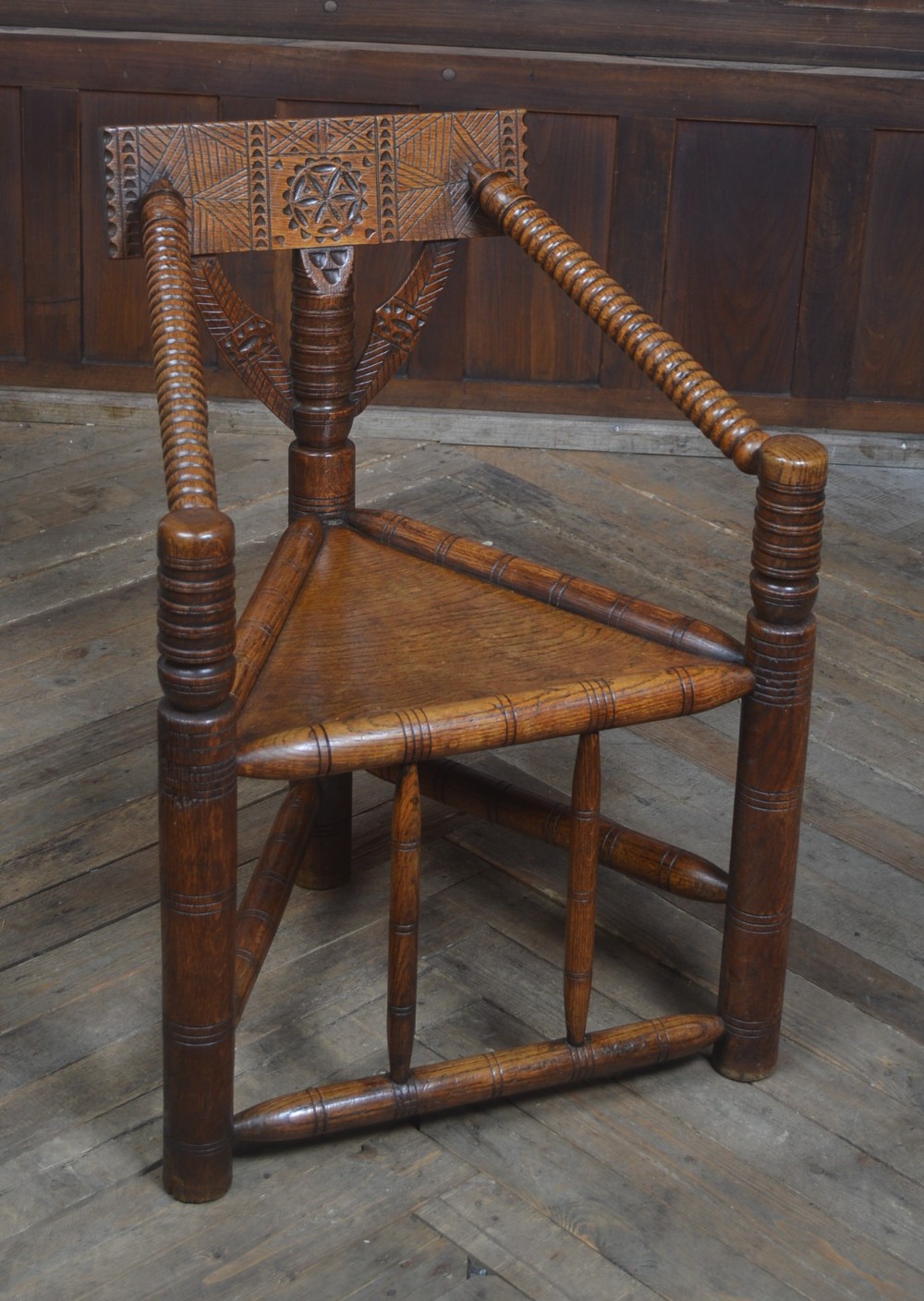 A carved oak Turners armchair featuring a triangular seat, three lathe-turned legs, bobbin-turned armrests, and geometric chip carving on the crest rail.