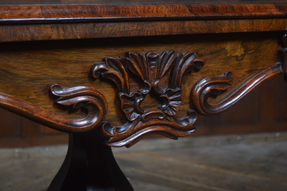 A Victorian rosewood fold-over card table featuring a carved frieze, central turned pedestal column, and scroll-footed quadform base on castors. View III