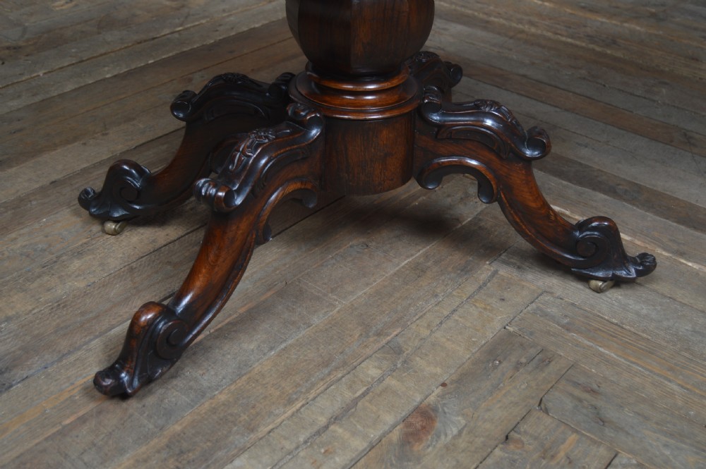 A Victorian rosewood fold-over card table featuring a carved frieze, central turned pedestal column, and scroll-footed quadform base on castors. View V