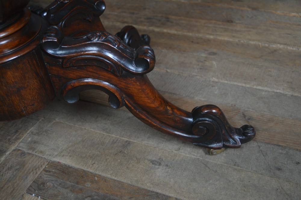 A Victorian rosewood fold-over card table featuring a carved frieze, central turned pedestal column, and scroll-footed quadform base on castors. View XVII