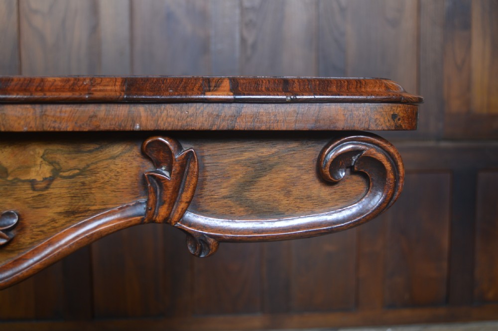 A Victorian rosewood fold-over card table featuring a carved frieze, central turned pedestal column, and scroll-footed quadform base on castors. View XVIII