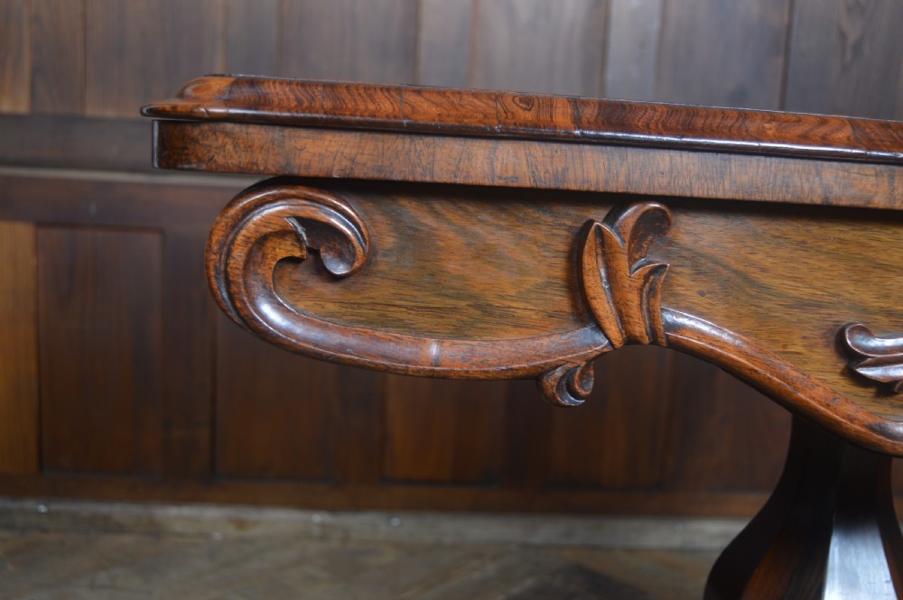 A Victorian rosewood fold-over card table featuring a carved frieze, central turned pedestal column, and scroll-footed quadform base on castors. View XIX