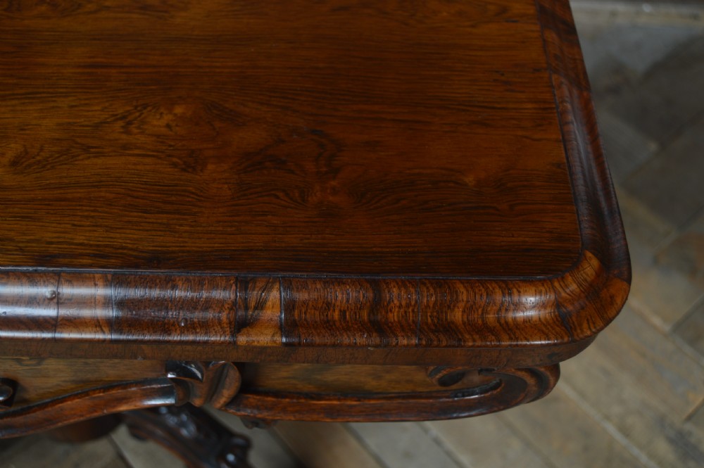 A Victorian rosewood fold-over card table featuring a carved frieze, central turned pedestal column, and scroll-footed quadform base on castors. View XXI