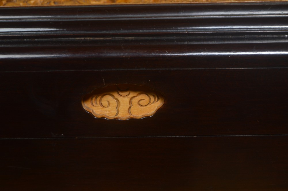 A rectangular dark lacquered storage trunk featuring intricate relief-carved light wood panels on the top, front, and sides depicting human figures and architectural structures, supported by carved corner columns and fitted with a central brass latch. View V