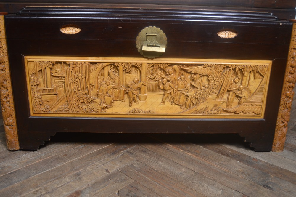 A rectangular dark lacquered storage trunk featuring intricate relief-carved light wood panels on the top, front, and sides depicting human figures and architectural structures, supported by carved corner columns and fitted with a central brass latch. View VII