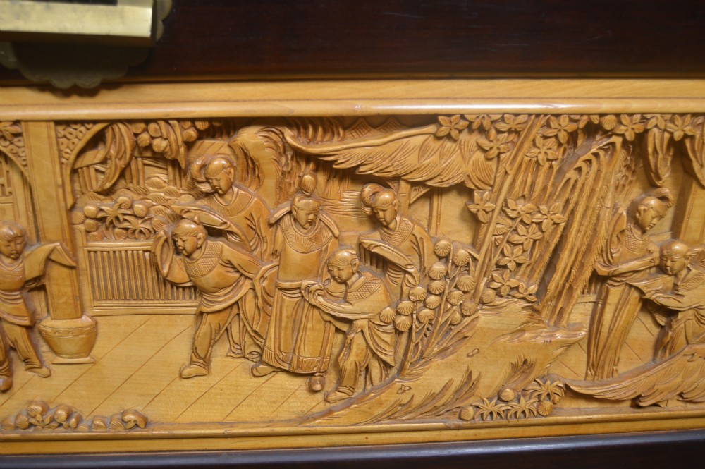A rectangular dark lacquered storage trunk featuring intricate relief-carved light wood panels on the top, front, and sides depicting human figures and architectural structures, supported by carved corner columns and fitted with a central brass latch. View VIII