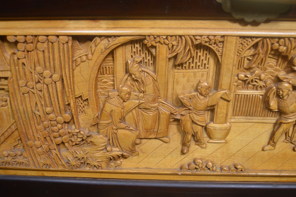 A rectangular dark lacquered storage trunk featuring intricate relief-carved light wood panels on the top, front, and sides depicting human figures and architectural structures, supported by carved corner columns and fitted with a central brass latch. View IX