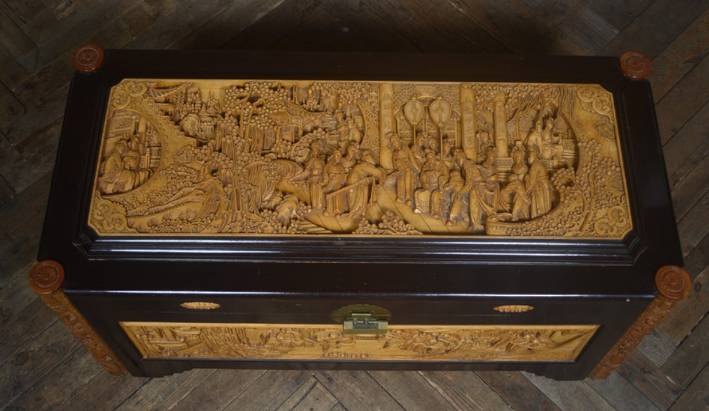 A rectangular dark lacquered storage trunk featuring intricate relief-carved light wood panels on the top, front, and sides depicting human figures and architectural structures, supported by carved corner columns and fitted with a central brass latch. View X