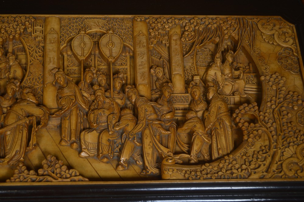 A rectangular dark lacquered storage trunk featuring intricate relief-carved light wood panels on the top, front, and sides depicting human figures and architectural structures, supported by carved corner columns and fitted with a central brass latch. View XI