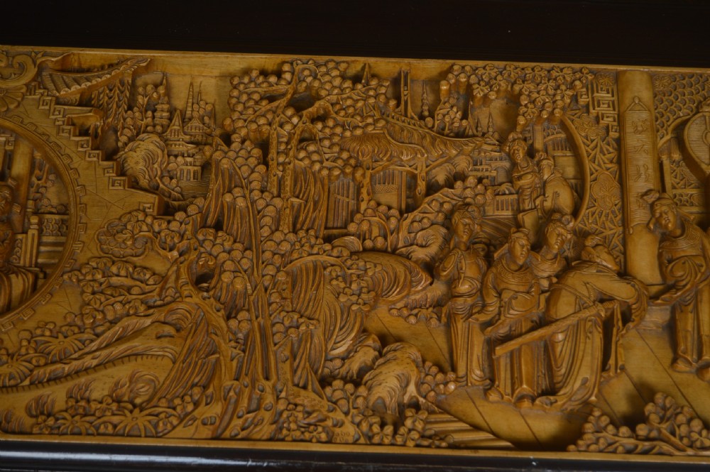 A rectangular dark lacquered storage trunk featuring intricate relief-carved light wood panels on the top, front, and sides depicting human figures and architectural structures, supported by carved corner columns and fitted with a central brass latch. View XII