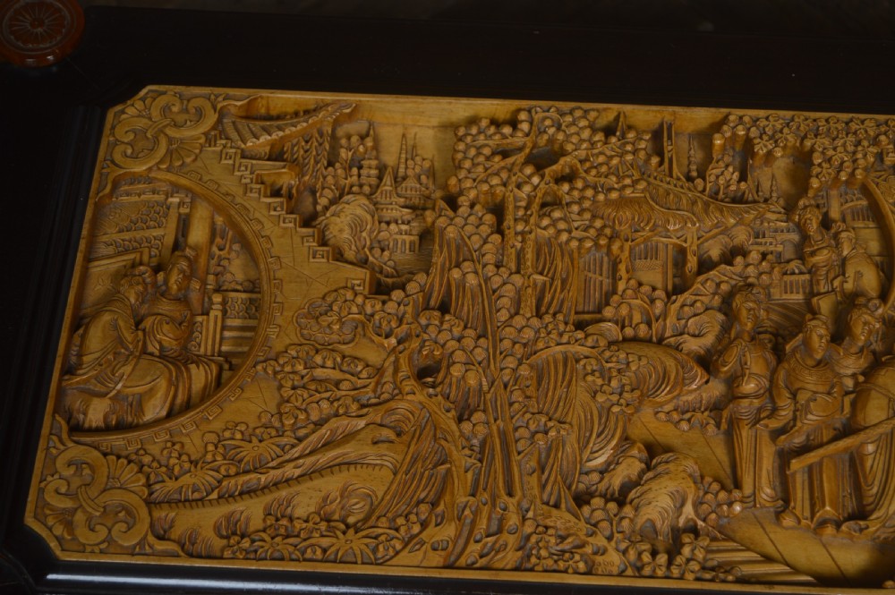 A rectangular dark lacquered storage trunk featuring intricate relief-carved light wood panels on the top, front, and sides depicting human figures and architectural structures, supported by carved corner columns and fitted with a central brass latch. View XIII