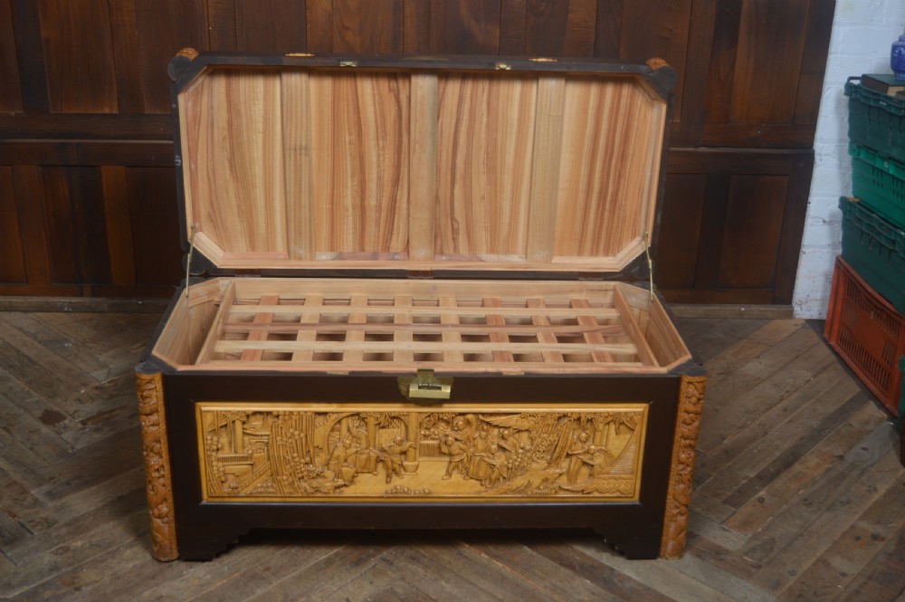 A rectangular dark lacquered storage trunk featuring intricate relief-carved light wood panels on the top, front, and sides depicting human figures and architectural structures, supported by carved corner columns and fitted with a central brass latch. View XIV
