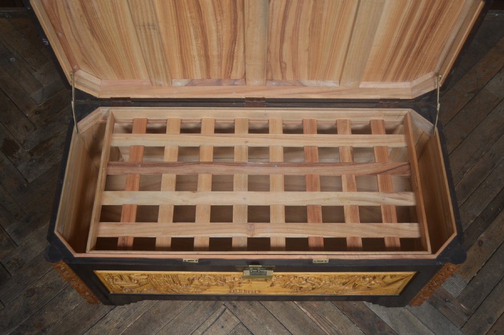 A rectangular dark lacquered storage trunk featuring intricate relief-carved light wood panels on the top, front, and sides depicting human figures and architectural structures, supported by carved corner columns and fitted with a central brass latch. View XV