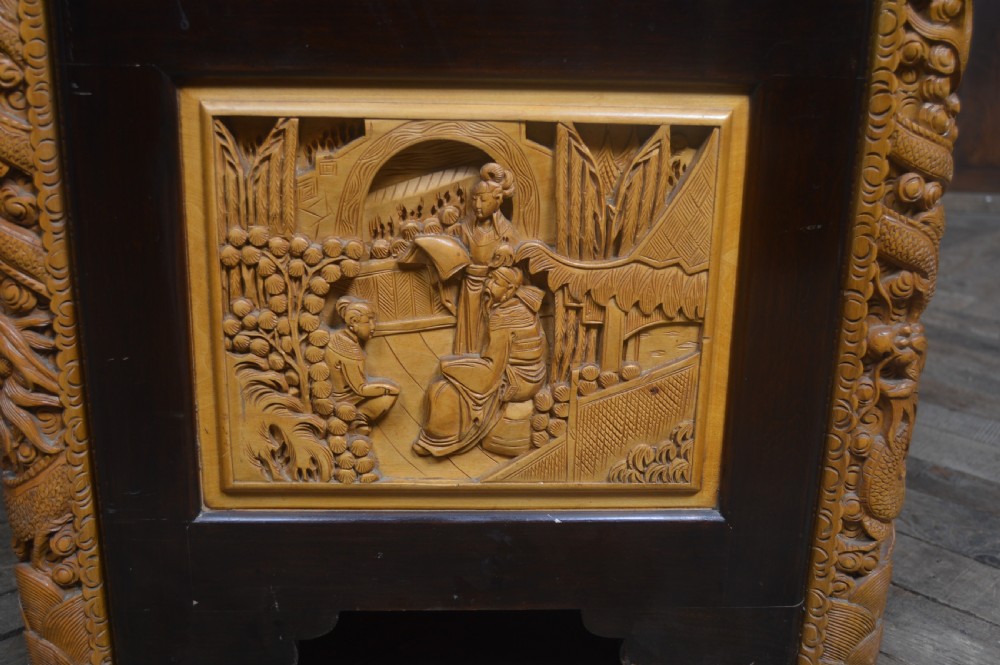 A rectangular dark lacquered storage trunk featuring intricate relief-carved light wood panels on the top, front, and sides depicting human figures and architectural structures, supported by carved corner columns and fitted with a central brass latch. View XVIII