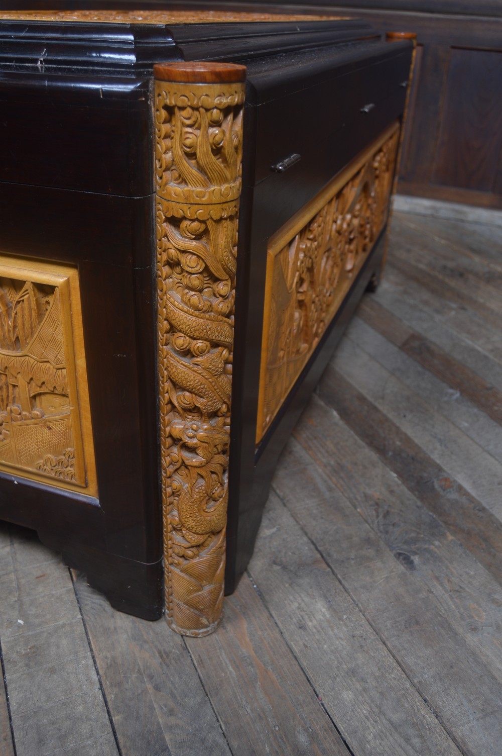 A rectangular dark lacquered storage trunk featuring intricate relief-carved light wood panels on the top, front, and sides depicting human figures and architectural structures, supported by carved corner columns and fitted with a central brass latch. View XIX
