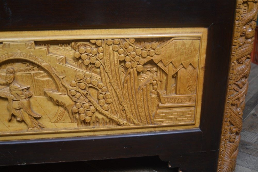 A rectangular dark lacquered storage trunk featuring intricate relief-carved light wood panels on the top, front, and sides depicting human figures and architectural structures, supported by carved corner columns and fitted with a central brass latch. View XXI