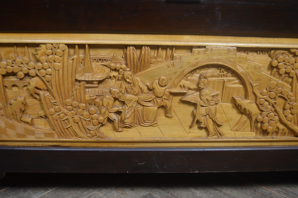 A rectangular dark lacquered storage trunk featuring intricate relief-carved light wood panels on the top, front, and sides depicting human figures and architectural structures, supported by carved corner columns and fitted with a central brass latch. View XXII
