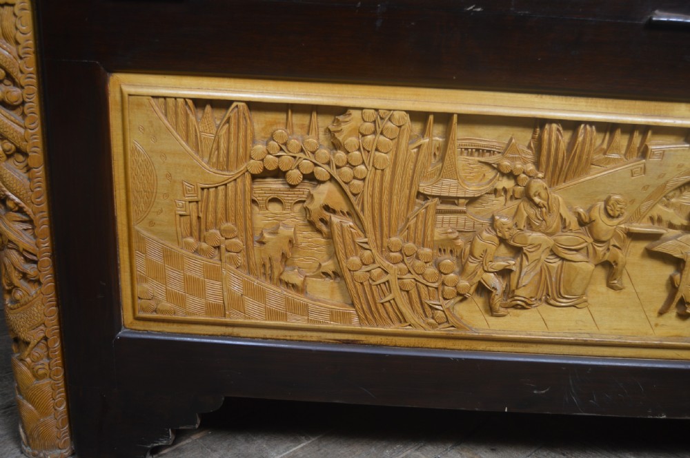 A rectangular dark lacquered storage trunk featuring intricate relief-carved light wood panels on the top, front, and sides depicting human figures and architectural structures, supported by carved corner columns and fitted with a central brass latch. View XXIII