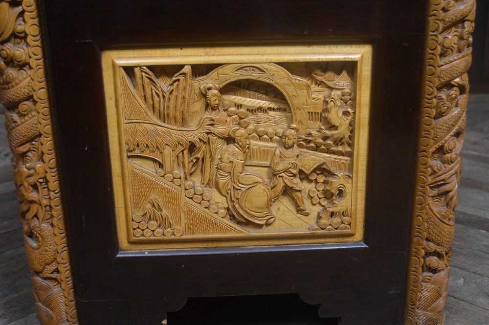 A rectangular dark lacquered storage trunk featuring intricate relief-carved light wood panels on the top, front, and sides depicting human figures and architectural structures, supported by carved corner columns and fitted with a central brass latch. View XXV