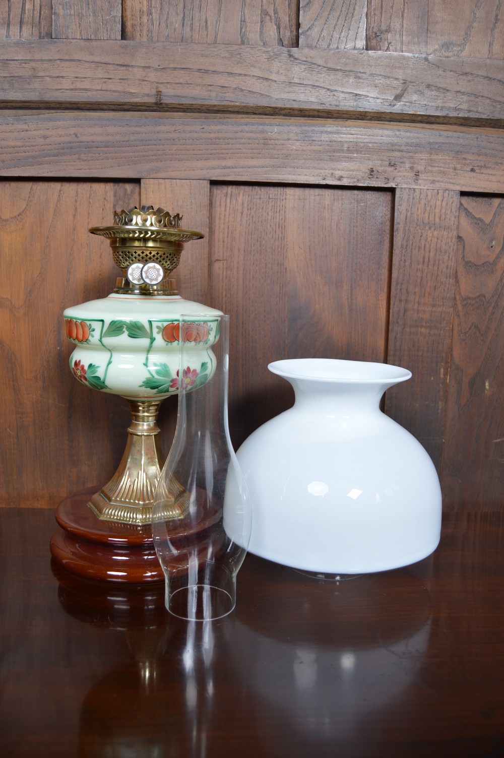 An antique Victorian paraffin oil lamp featuring a hand-painted floral glass reservoir, a brass fluted stem, a white opaline shade, and a circular wooden plinth base. View X