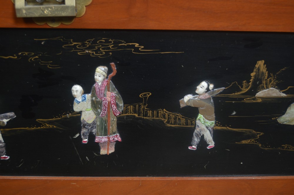 A Chinese camphor wood storage chest featuring a reddish-brown frame, black lacquer panels with figurative inlays, and a brass front clasp. View V