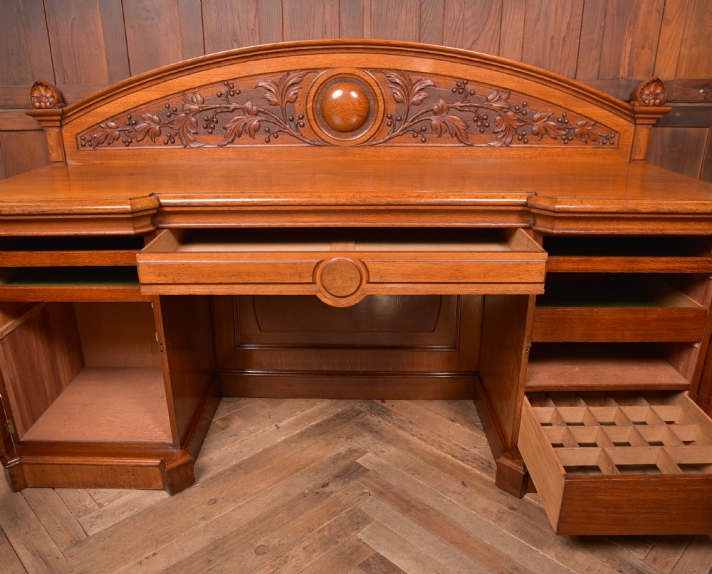 A Victorian oak pedestal sideboard featuring an arched carved backboard with foliage details, twin cupboard pedestals, and a recessed central section. View XIV