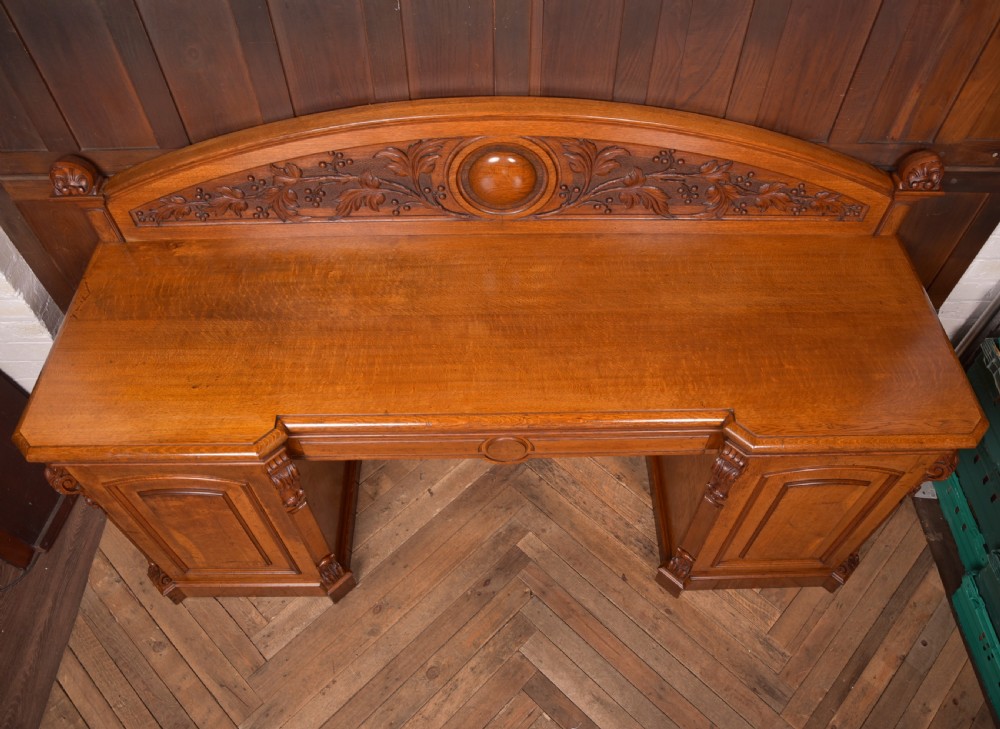 A Victorian oak pedestal sideboard featuring an arched carved backboard with foliage details, twin cupboard pedestals, and a recessed central section. View XV