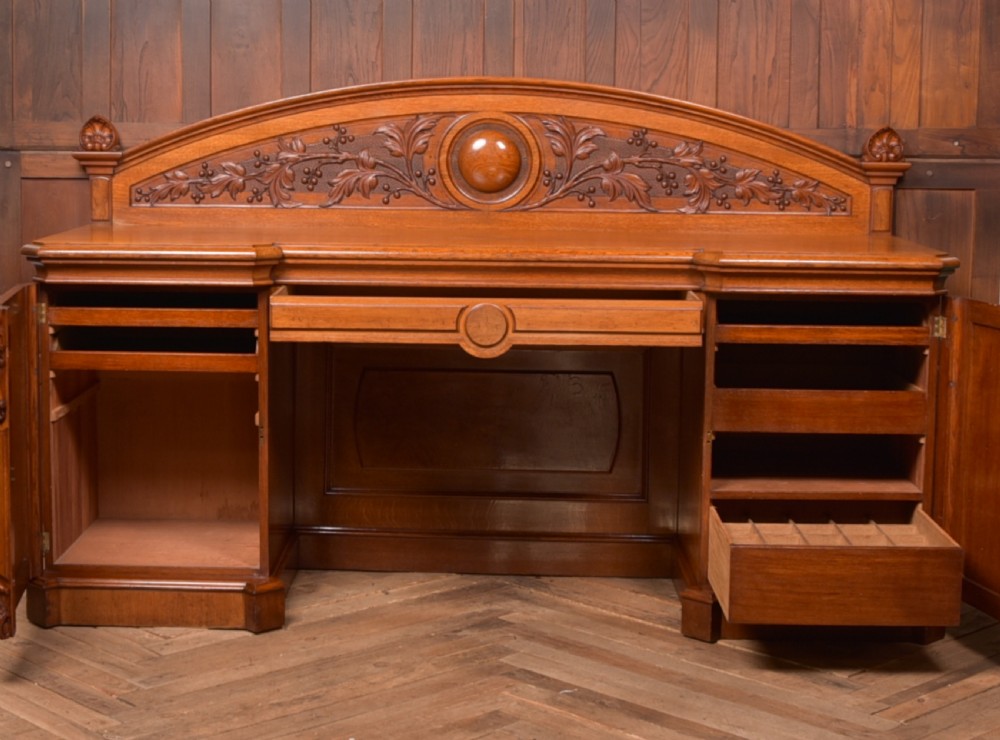 A Victorian oak pedestal sideboard featuring an arched carved backboard with foliage details, twin cupboard pedestals, and a recessed central section. View XVII