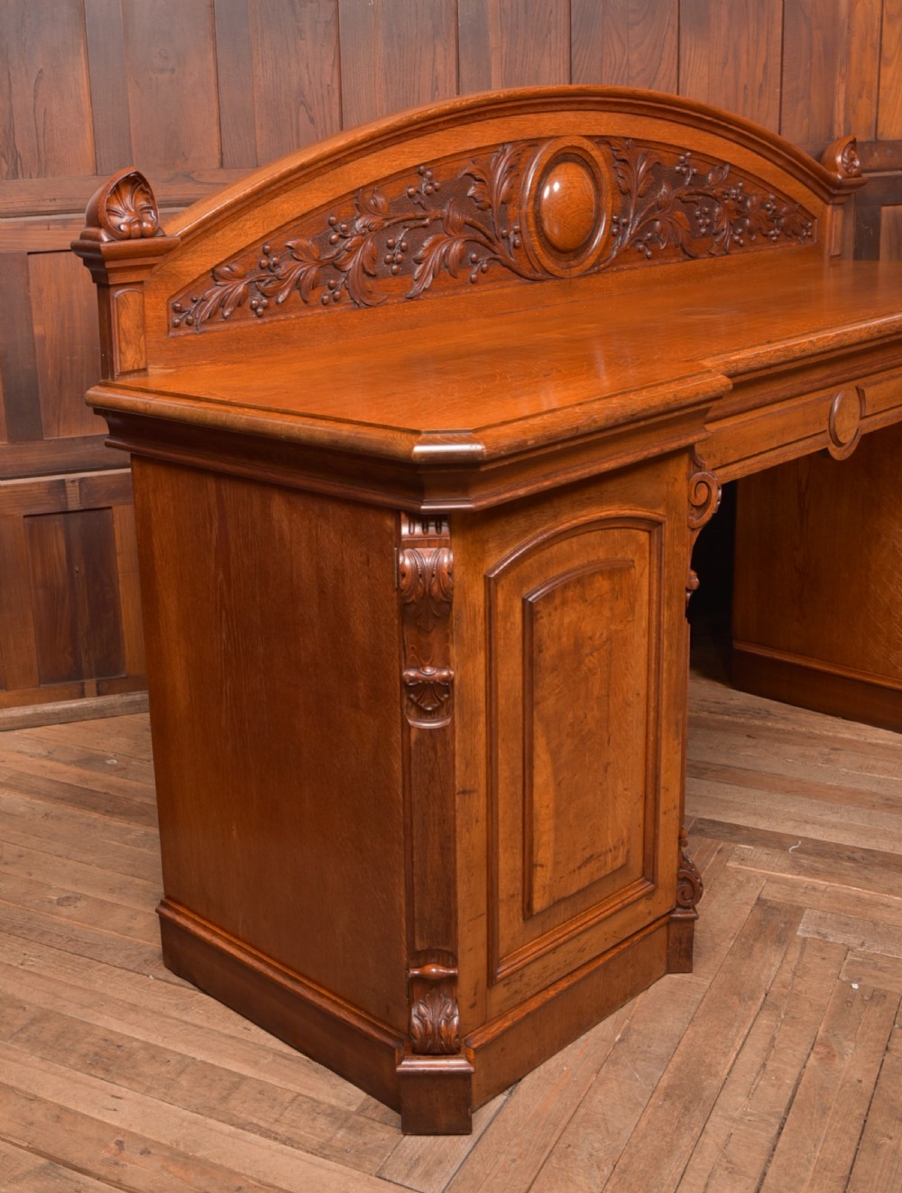 A Victorian oak pedestal sideboard featuring an arched carved backboard with foliage details, twin cupboard pedestals, and a recessed central section. View XVIII