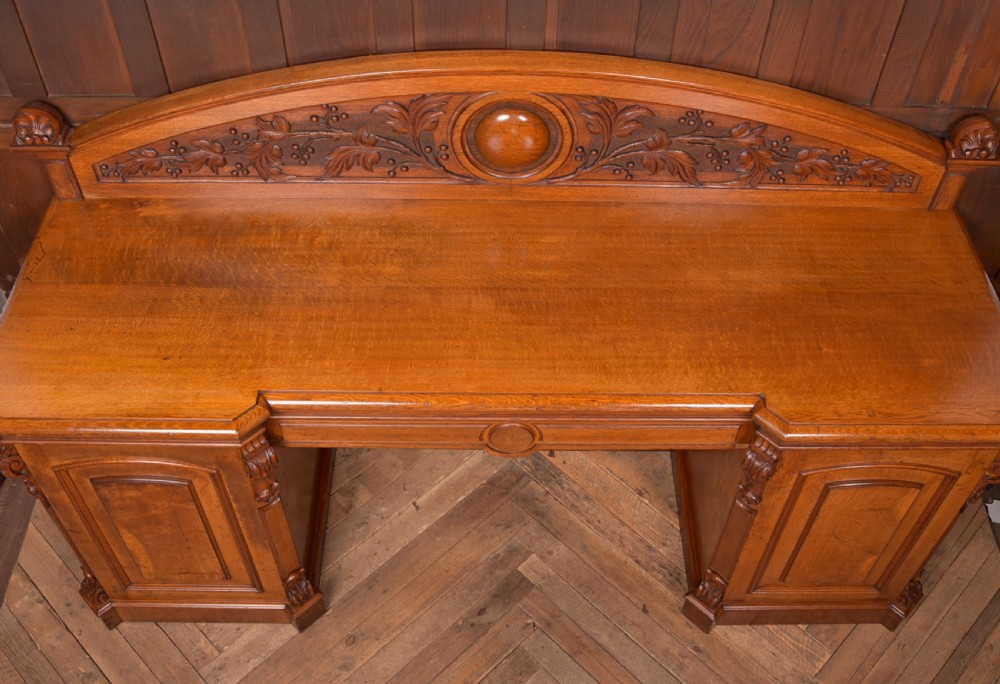 A Victorian oak pedestal sideboard featuring an arched carved backboard with foliage details, twin cupboard pedestals, and a recessed central section. View XIX