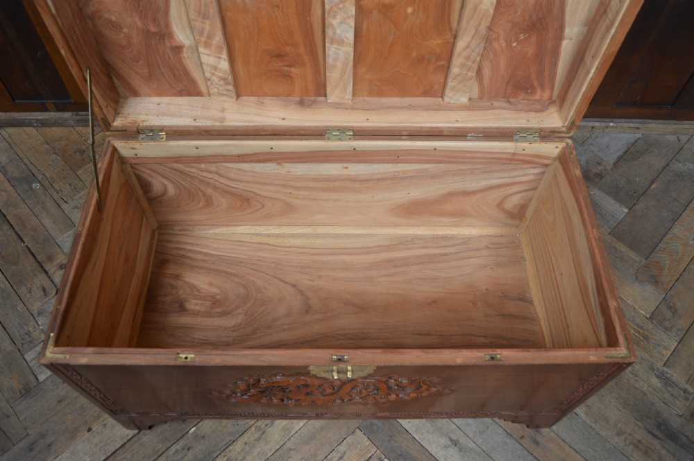 A carved warm brown camphor wood chest featuring a relief dragon motif on the front panel and a decorative circular brass clasp. View X