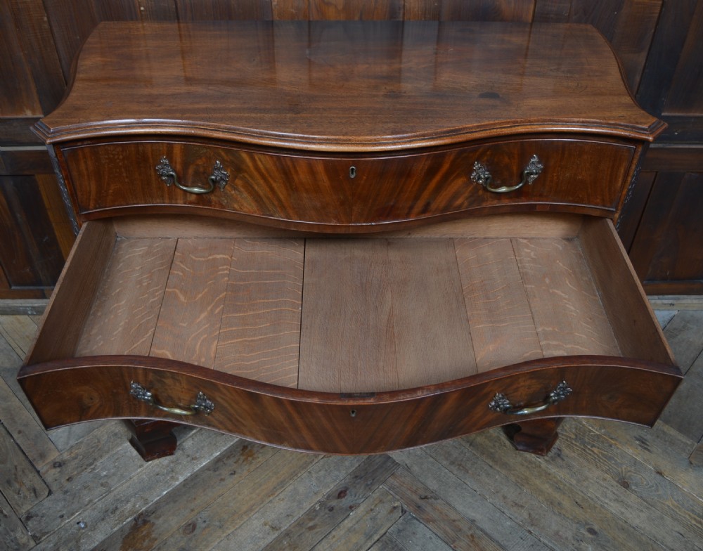 An 18th-century Georgian flame mahogany serpentine chest of drawers featuring four graduated drawers with rococo brass handles and ogee bracket feet. View X