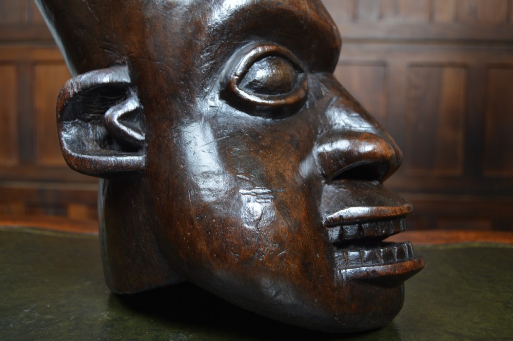 A hand-carved dark wood African Bamum helmet mask featuring exaggerated facial features, almond-shaped eyes, an open mouth with visible teeth, and a crest atop the head. View IX
