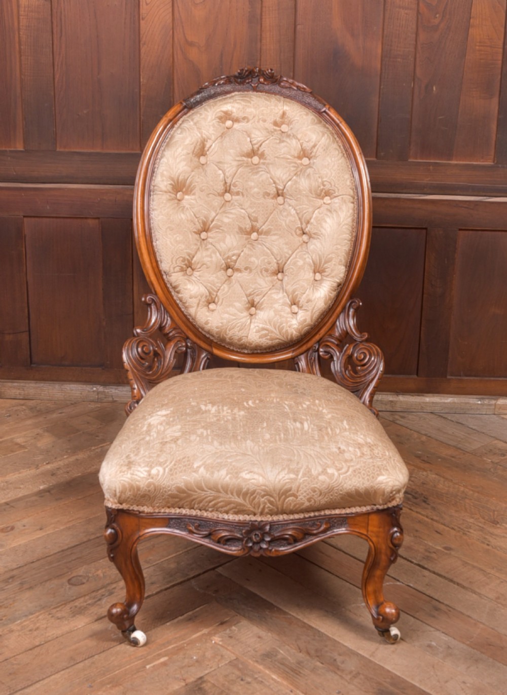 A Victorian carved walnut nursing chair with a button-tufted oval backrest, champagne fabric upholstery, carved cabriole legs, and ceramic castors.