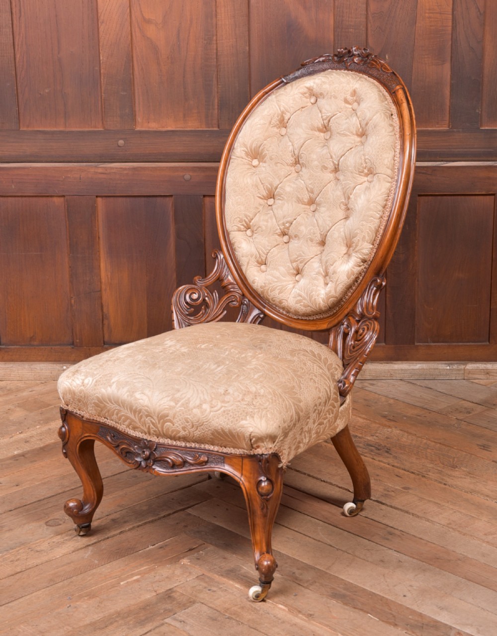 A Victorian carved walnut nursing chair with a button-tufted oval backrest, champagne fabric upholstery, carved cabriole legs, and ceramic castors. View II