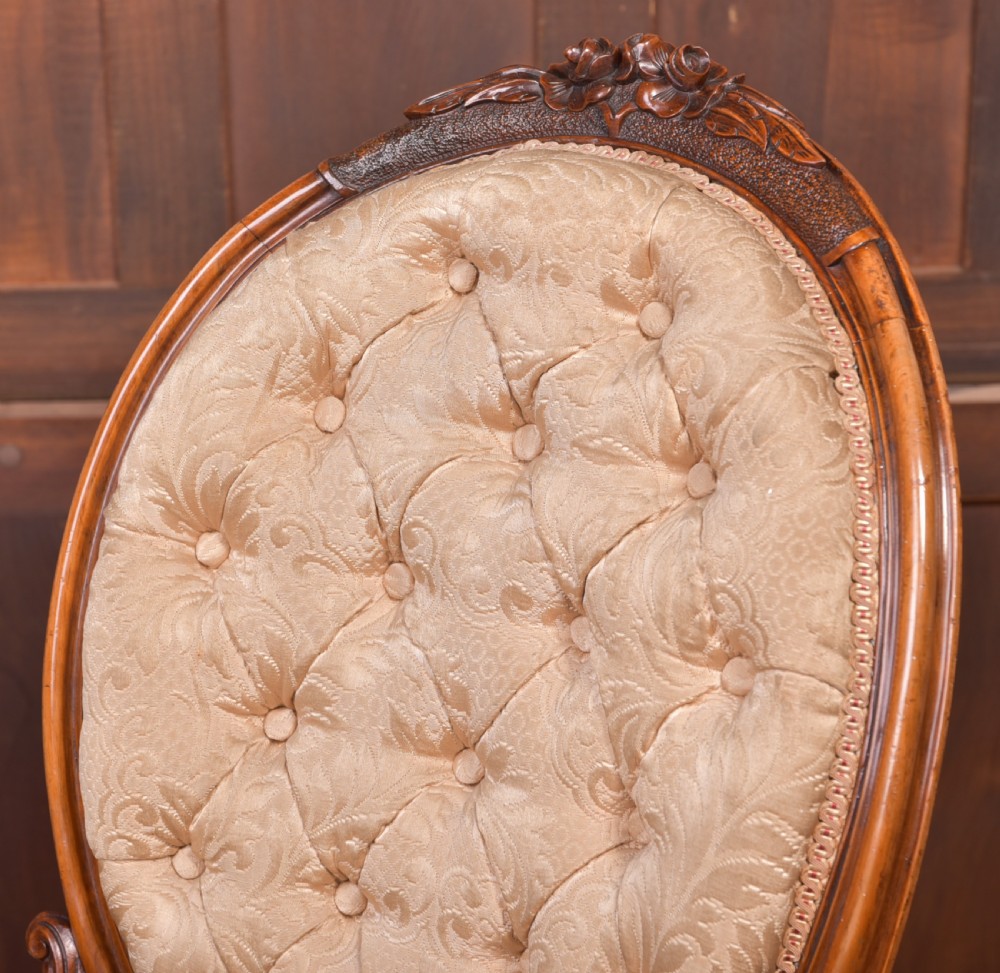 A Victorian carved walnut nursing chair with a button-tufted oval backrest, champagne fabric upholstery, carved cabriole legs, and ceramic castors. View III