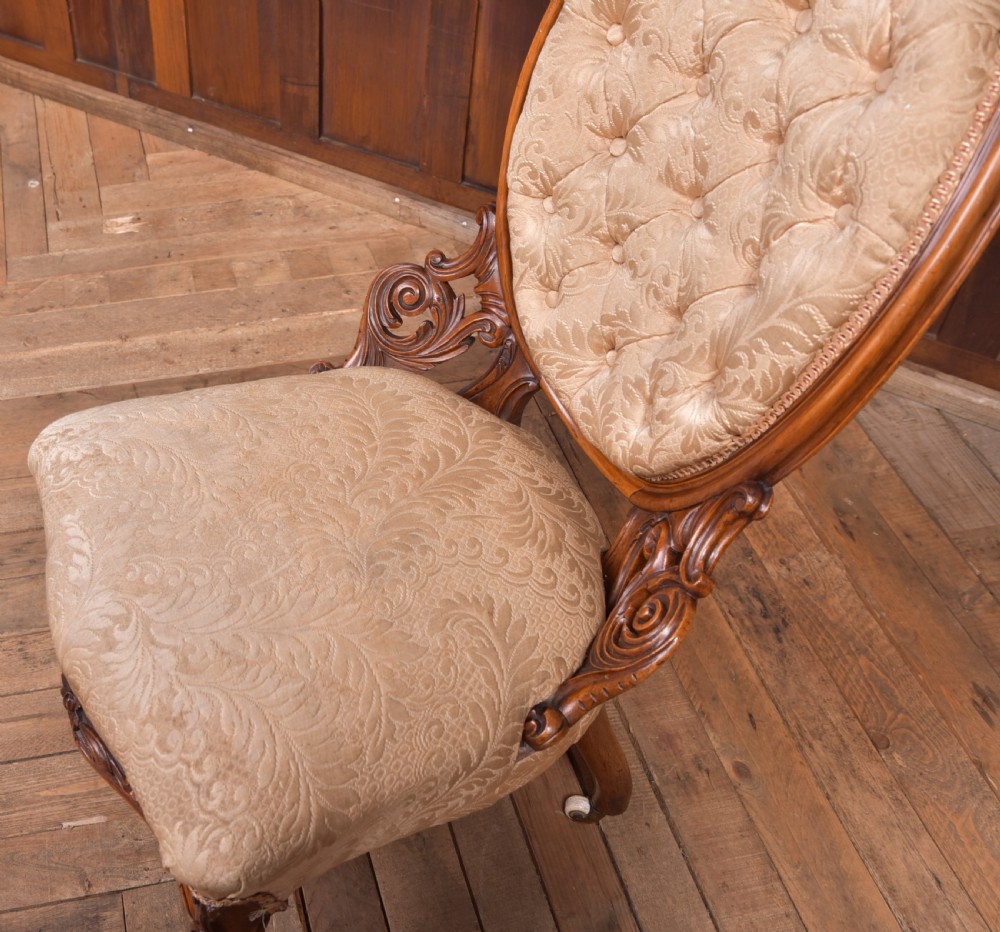 A Victorian carved walnut nursing chair with a button-tufted oval backrest, champagne fabric upholstery, carved cabriole legs, and ceramic castors. View V