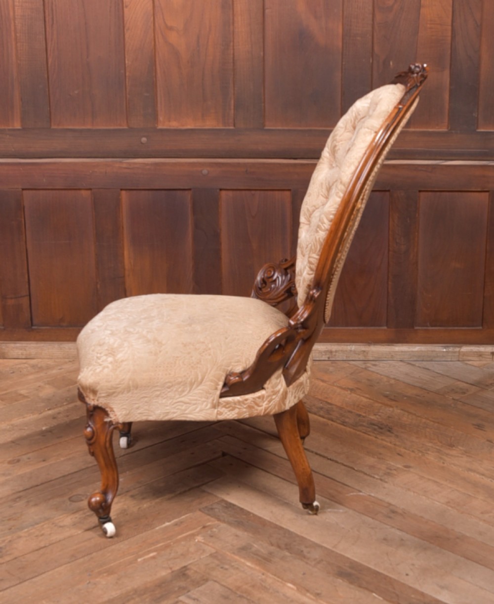 A Victorian carved walnut nursing chair with a button-tufted oval backrest, champagne fabric upholstery, carved cabriole legs, and ceramic castors. View VI