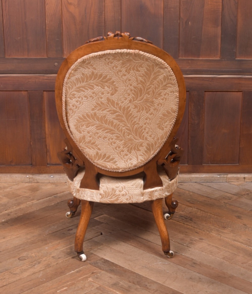 A Victorian carved walnut nursing chair with a button-tufted oval backrest, champagne fabric upholstery, carved cabriole legs, and ceramic castors. View VII