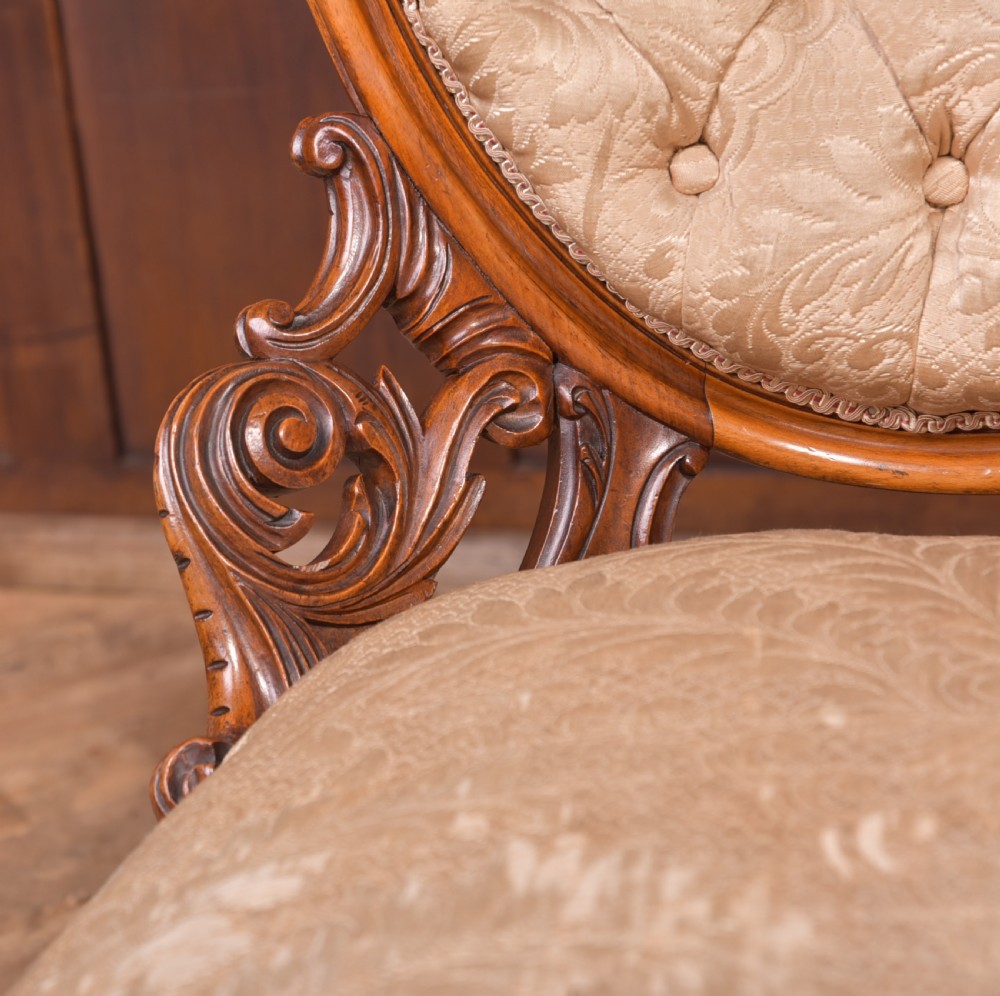 A Victorian carved walnut nursing chair with a button-tufted oval backrest, champagne fabric upholstery, carved cabriole legs, and ceramic castors. View X